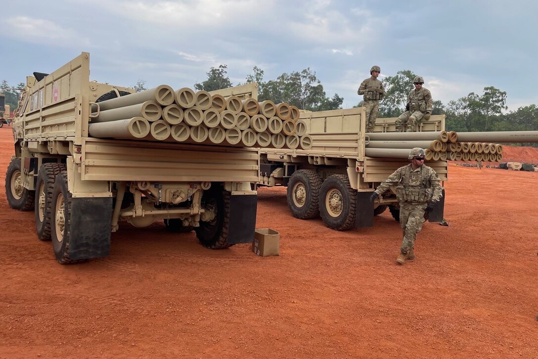Soldiers in uniform are photographed near trucks carrying stacks of pipe.