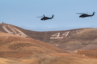Two helicopters fly over rolling brown hills with military insignia adorning them.