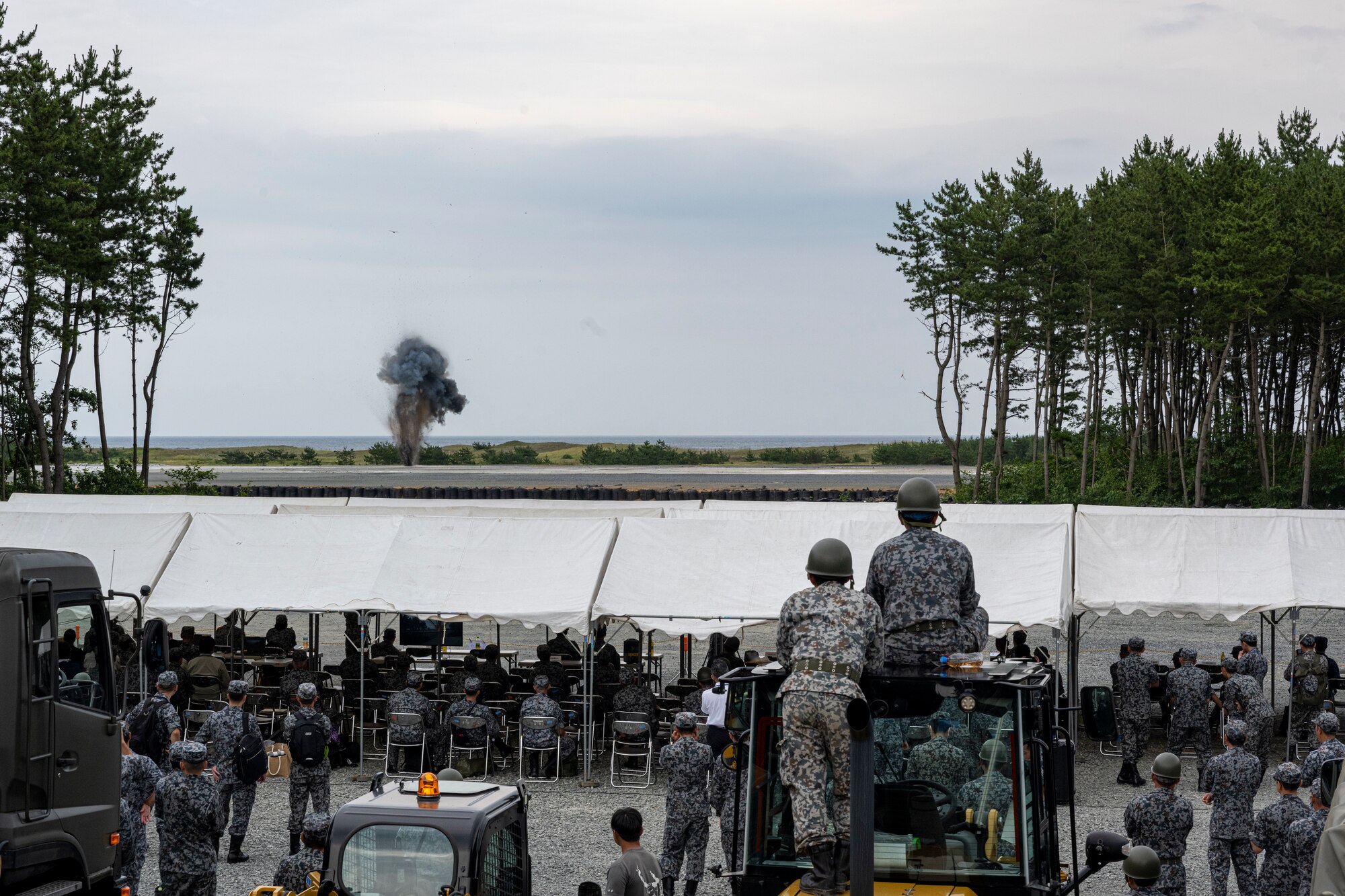 U.S. Air Force Airmen assigned to the 18th Civil Engineer Group and Japan Air Self-Defense Force Airmen assigned to the Western Air Civil Engineering Group watch as explosives explode on the rapid airfield damage recovery pad to simulate damage to a runway during a bilateral live fire exercise.