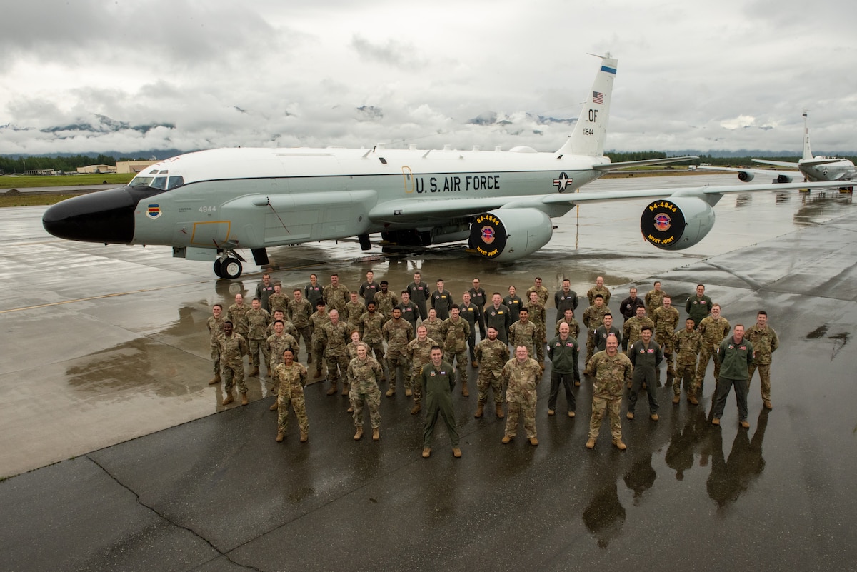 People in camouflage uniforms and flight suits pose in front of an aircraft on a gray, cloudy day