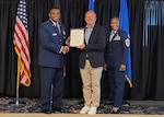 Dragan Civric stands in front of flags and flanked by Air Force Brig. Gen. Otis Jones and Air Force Chief Master Sgt. Charmaine Kelley as they hold an award