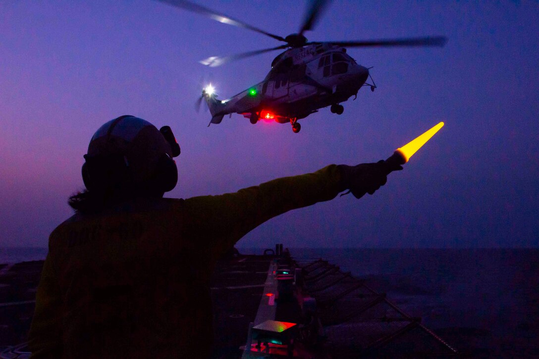 A sailor standing on the deck of a ship uses a light wand to signal toward an airborne helicopter at night.