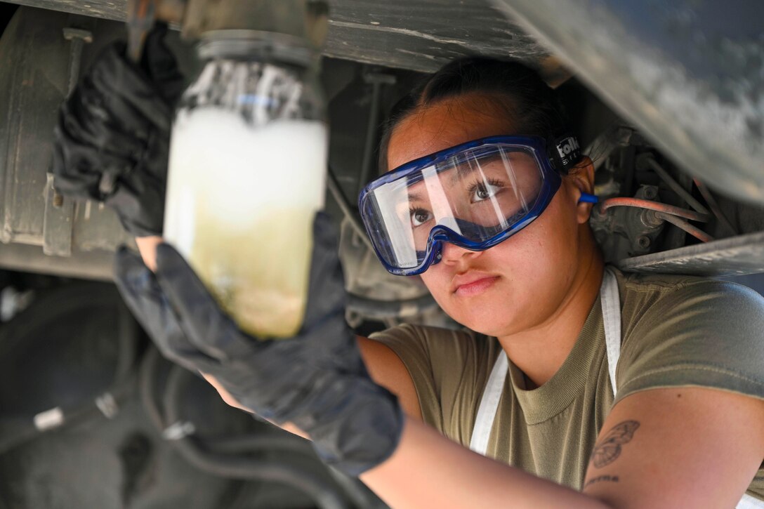 An airman wearing protective gear puts fuel in a jar while kneeling under an aircraft.