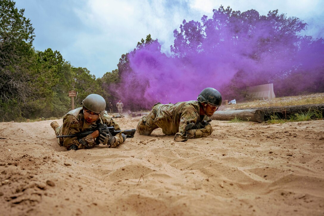 Airmen wearing tactical gear and carrying weapons crawl in sand under clouds of purple smoke.