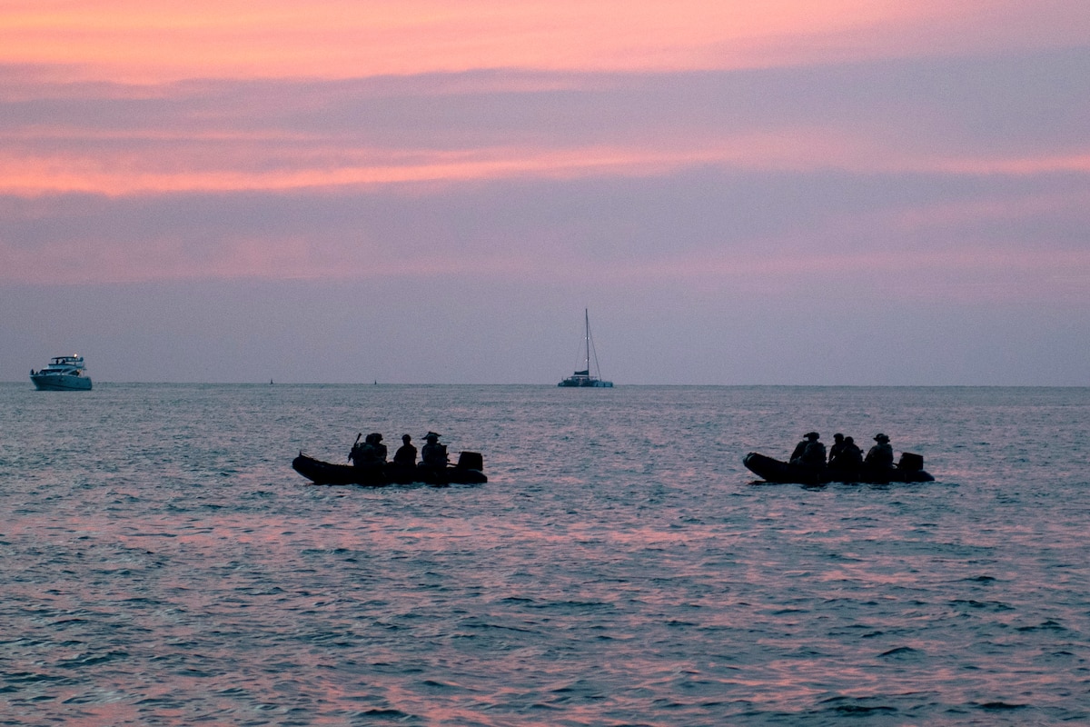 U.S. and Colombian forces in two small rubber boats transit a body of water with two ships in the background under a pinkish sky.