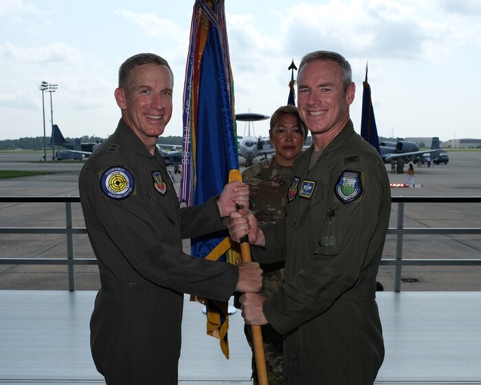 photo of three uniformed U.S. Air Force Airmen stand on a stand, two in the foreground holding a unit flag, while the third Airman stands at attention in the background are two A-10Cs and an airborne warning and control system, or AWACS, aircraft on Hurlburt Field flightline.
