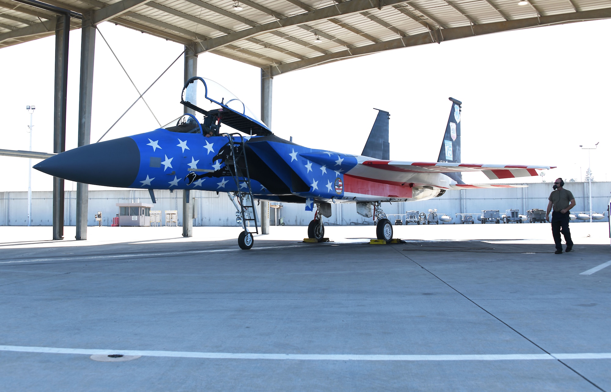 An F-15C fighter aircraft with a red, white, and blue paint scheme is parked under an aircraft bay. An airman stands to the right side of the aircraft.