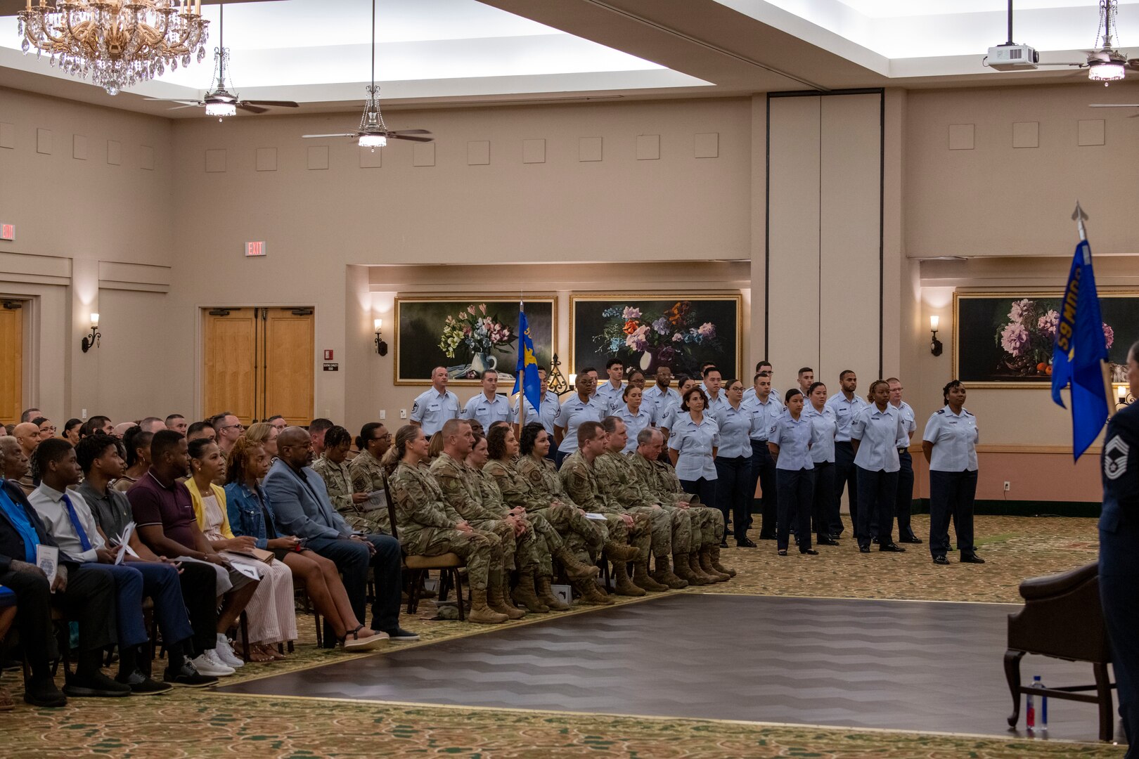 Audience sits at ceremony in foreground, military formation stands in background