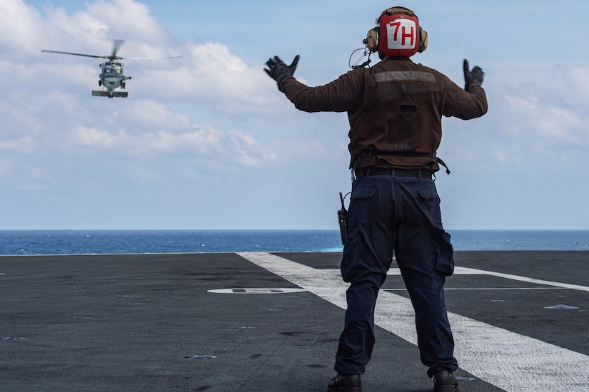 A sailor signals to an airborne helicopter from a ship at sea.
