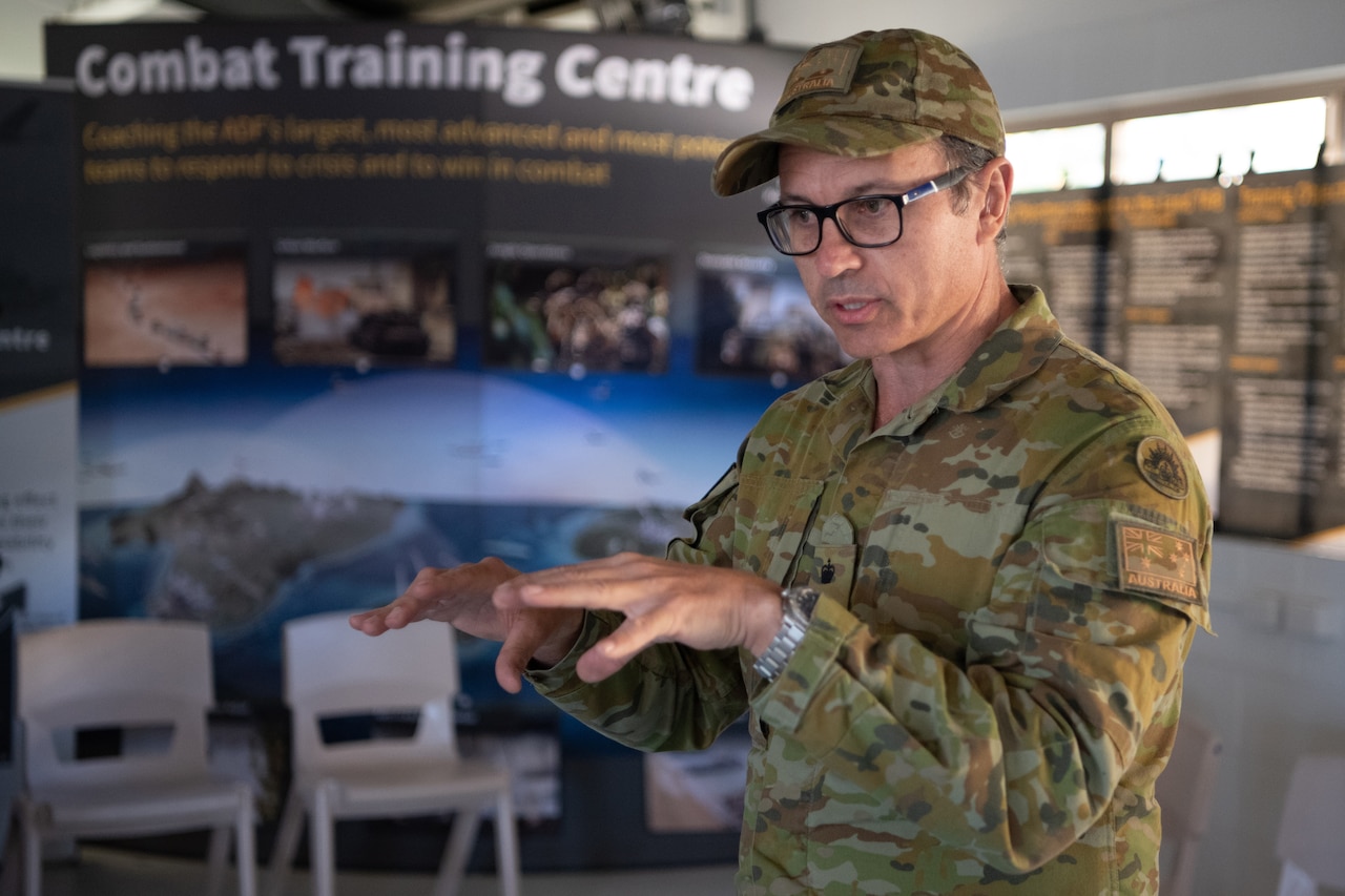 A man in a military uniform gestures with his hands; behind him is a sign bearing the words Combat Training Centre.