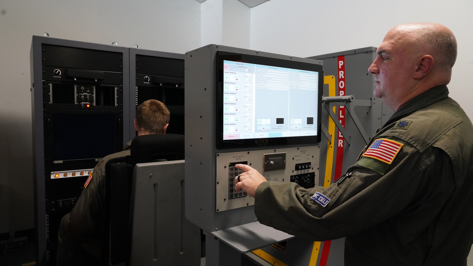 One male Airman stands behind the new weather officer static trainer to run the equipment while another sits in the seat.