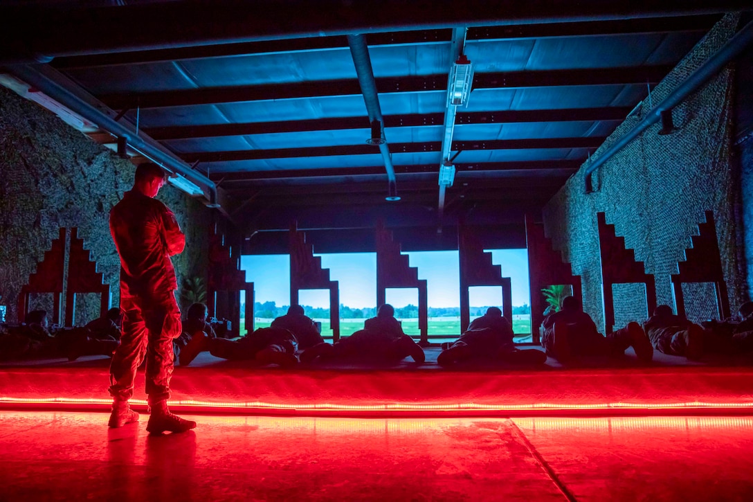 Soldiers lie on the ground in a dark room  over looking a shooting range as another soldier watches from behind.