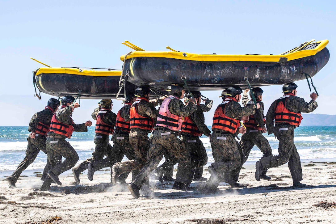 Two groups of sailors run on sand with two inflatable boats on their heads.