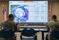 two men in army uniform sitting at a table talking in front of a large screen