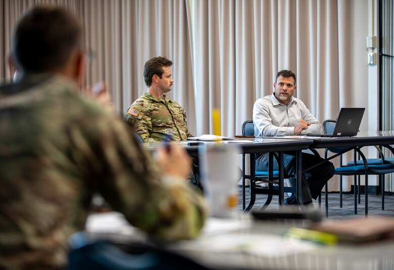 Men sit at tables while talking, two in army uniform.