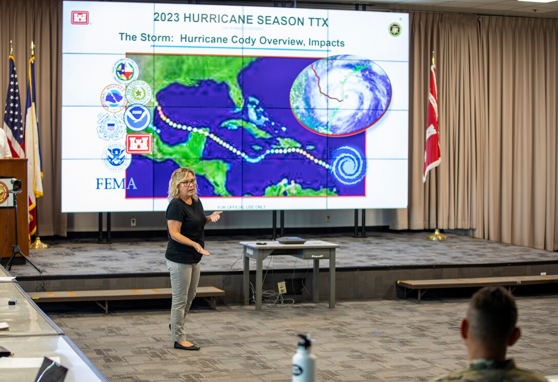 Woman is standing in front of a large screen speaking to other people