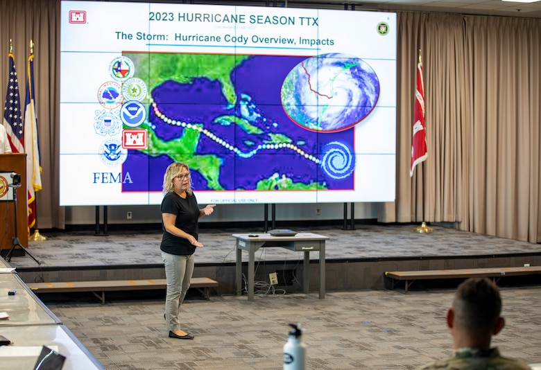 Woman is standing in front of a large screen speaking to other people