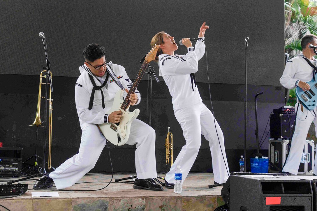A sailor plays guitar as another sings on a stage during a performance.