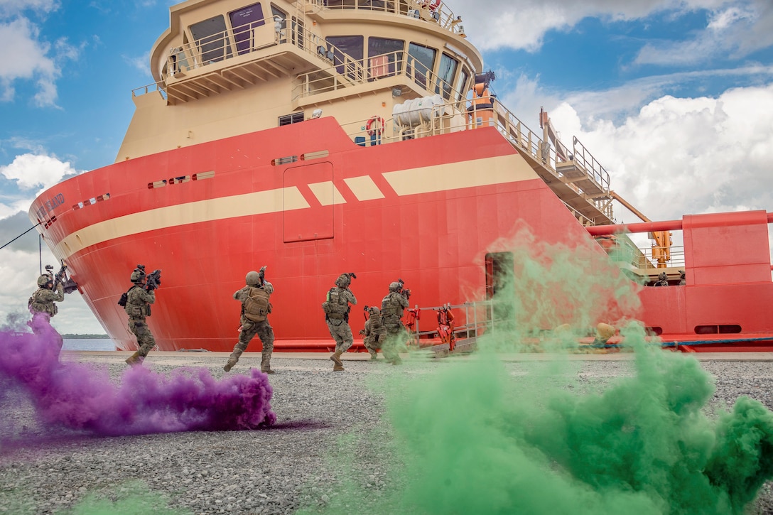 Troops aim weapons while boarding a moored ship surrounded by clouds of green and purple smoke.