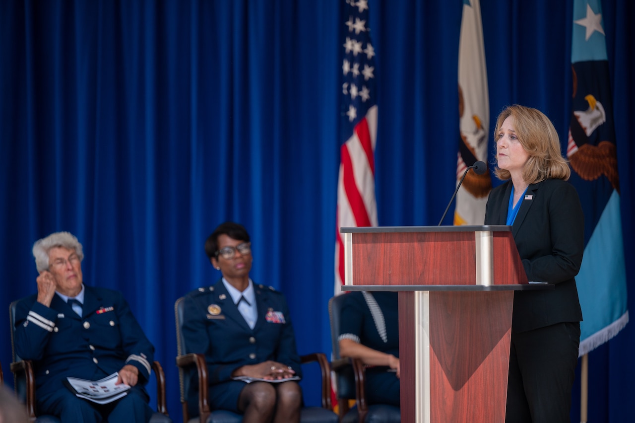 A woman talks at a lectern while others sit in chairs behind her.