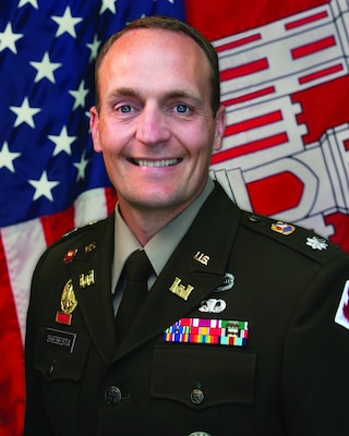 White male in Army greens with metals on stands in front of American flag - red, white and blue, and an Army Corps of Engineers flag - red and white castle.