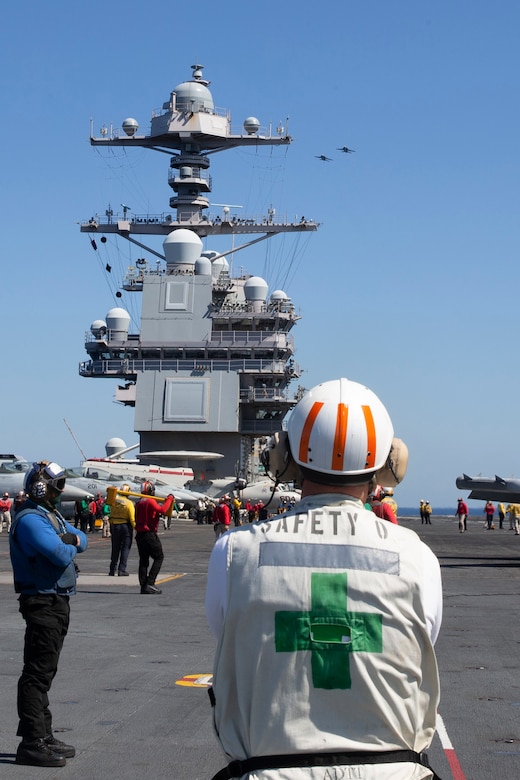 A sailor wearing white safety uniform stands on the deck of a ship with the mast up ahead and other sailors standing around.