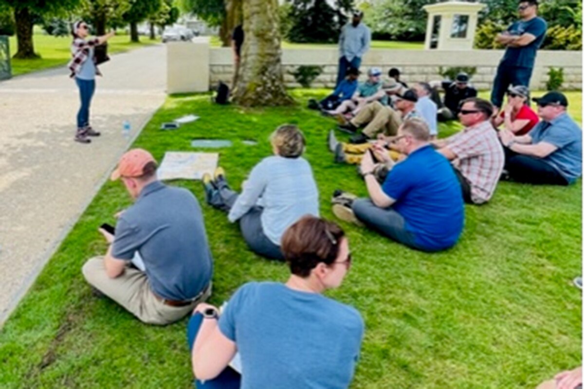 A group of people sit and stand on the grass under a tree, as another woman stands on a sidewalk in front of the group.