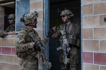 Two soldiers in battle gear on patrol bump fists in a doorway, as a third leans out a window.