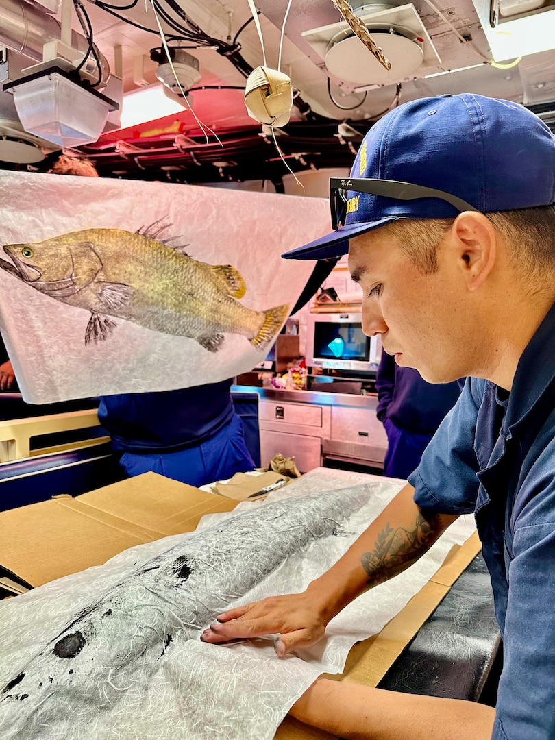 Petty Officer 1st Class Ikaika Ruiz presses a wahoo with Japanese rice paper for a Gyotaku print while aboard the USCGC Oliver Henry (WPC 1140) while underway off the Republic of Palau on April 1, 2023. His barramundi print can be seen in the background. Originally from Hawaii, Ruiz is an engineer aboard the ship specializing as an electrician. (U.S. Coast Guard photo by Chief Warrant Officer Sara Muir)