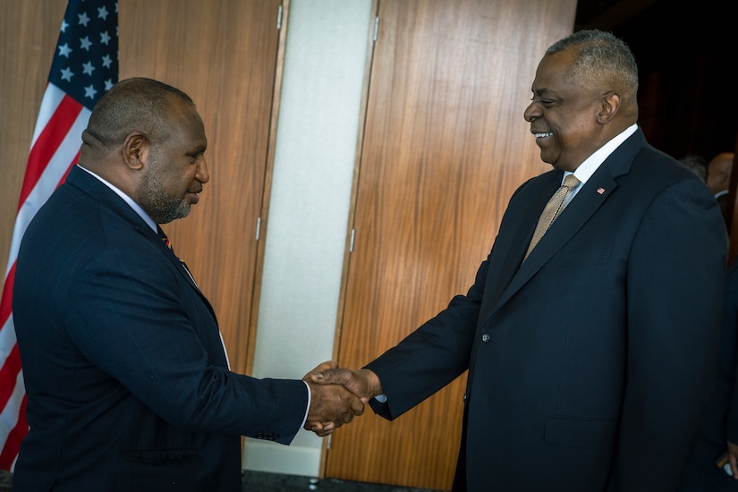 Two men shake hands with an American flag in the background.