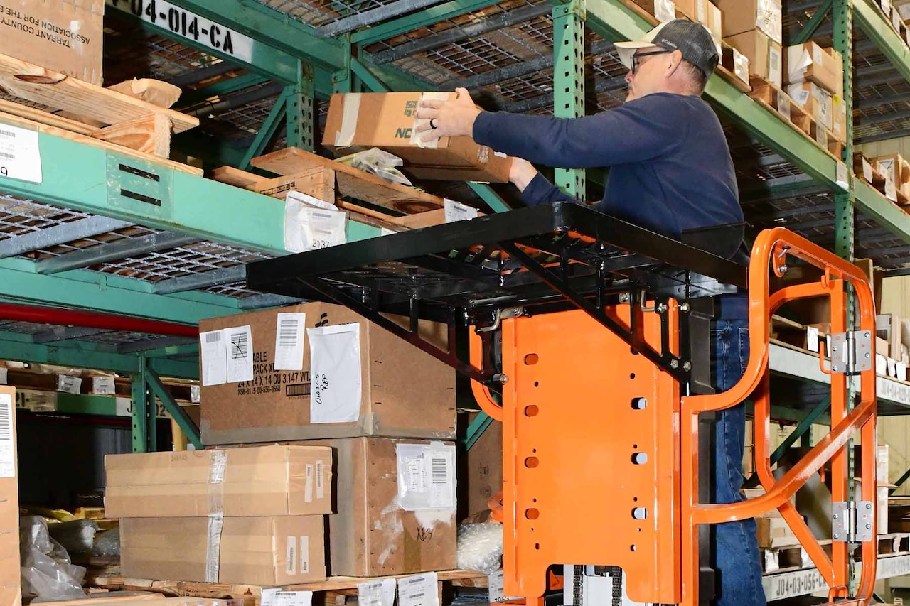 A man, standing in the cart of a lift, grabs an item from a shelf inside a warehouse.
