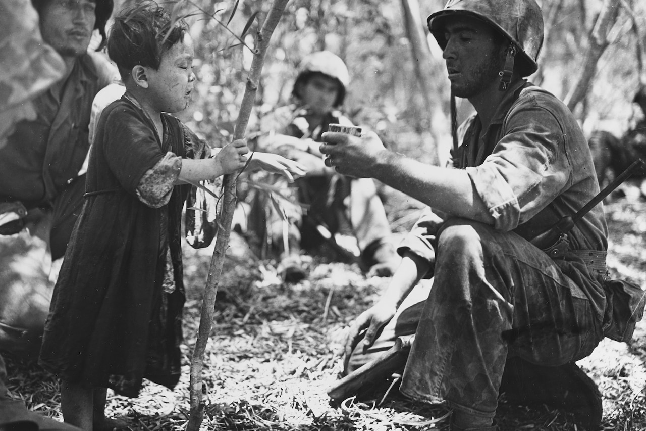 A man gives a small child a cup while in the woods.