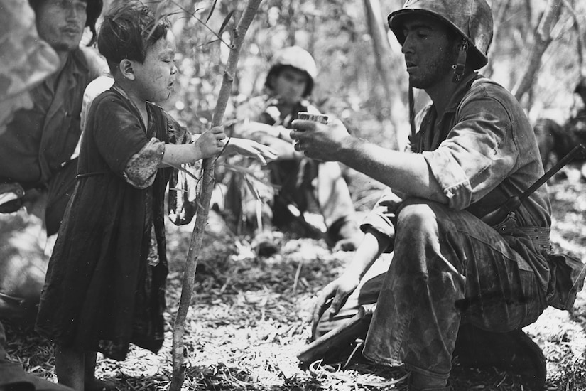A man gives a small child a cup while in the woods.