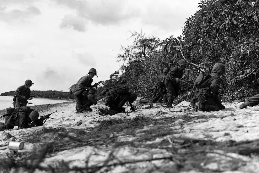 Men with guns rush onto the shore of a beach, heading for brush. A few are on their knees.