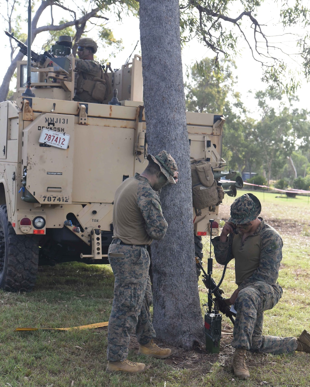 A Marine stands next to a tree watching another Marine work a radio.