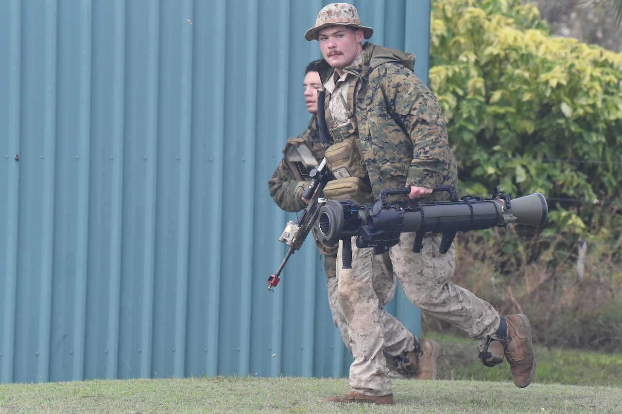 Two Marines carry their weapons as they run past a building.