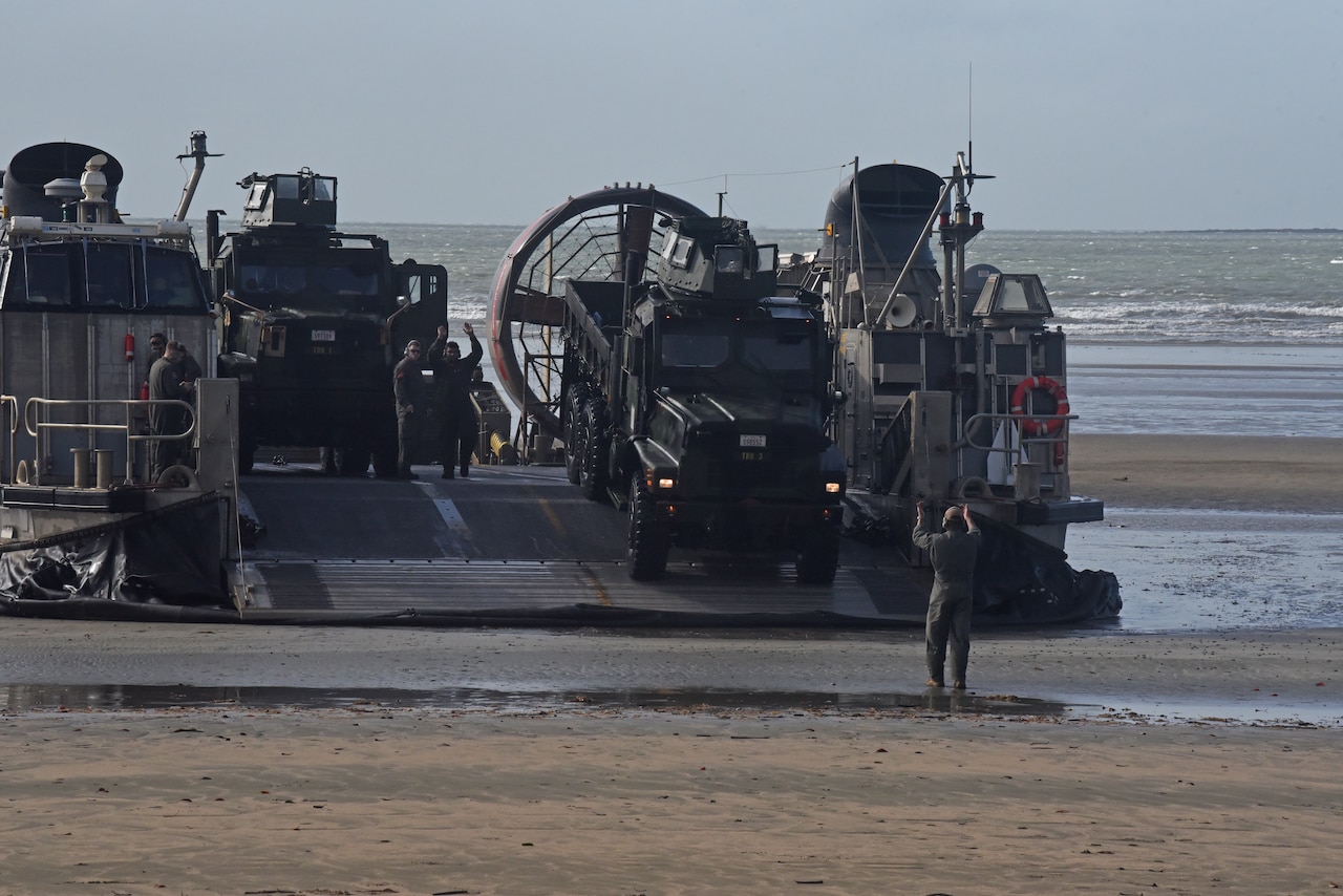 A service member guides a military vehicle off the ramp of a landing craft.