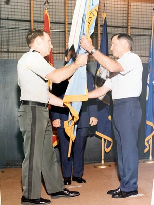 Photo is of a man in military uniform passing an organization flag to another man in military uniform, signifying a transition in leadership
