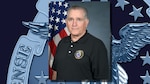 Photo is of a caucasian man in black shirt with U.S. flag over his right shoulder with a government seal in background