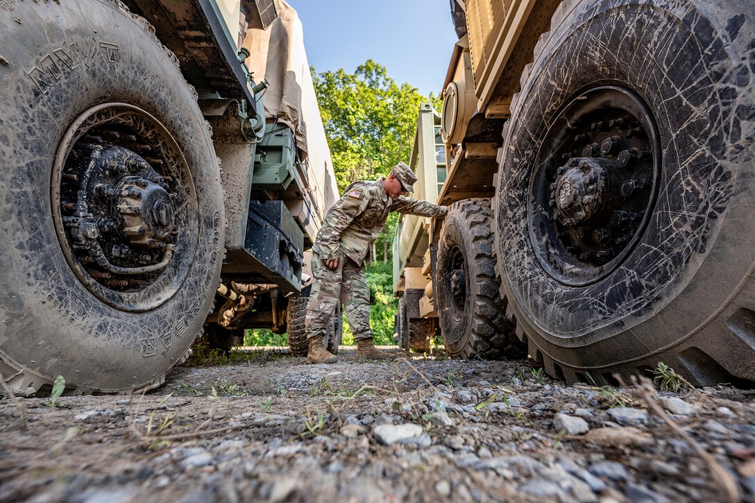 A service member stands between two military vehicles and checks a tire.
