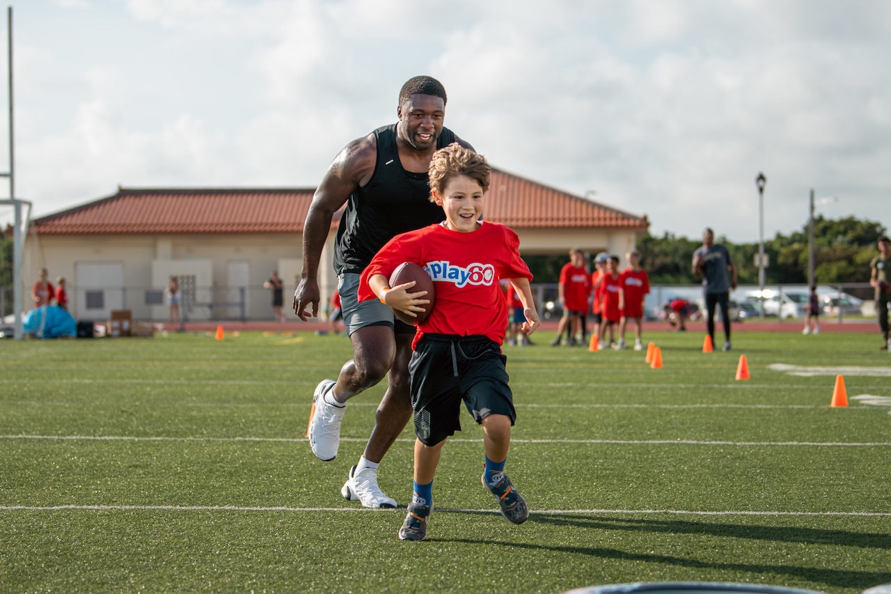 A smiling man runs after a smiling boy with a football.