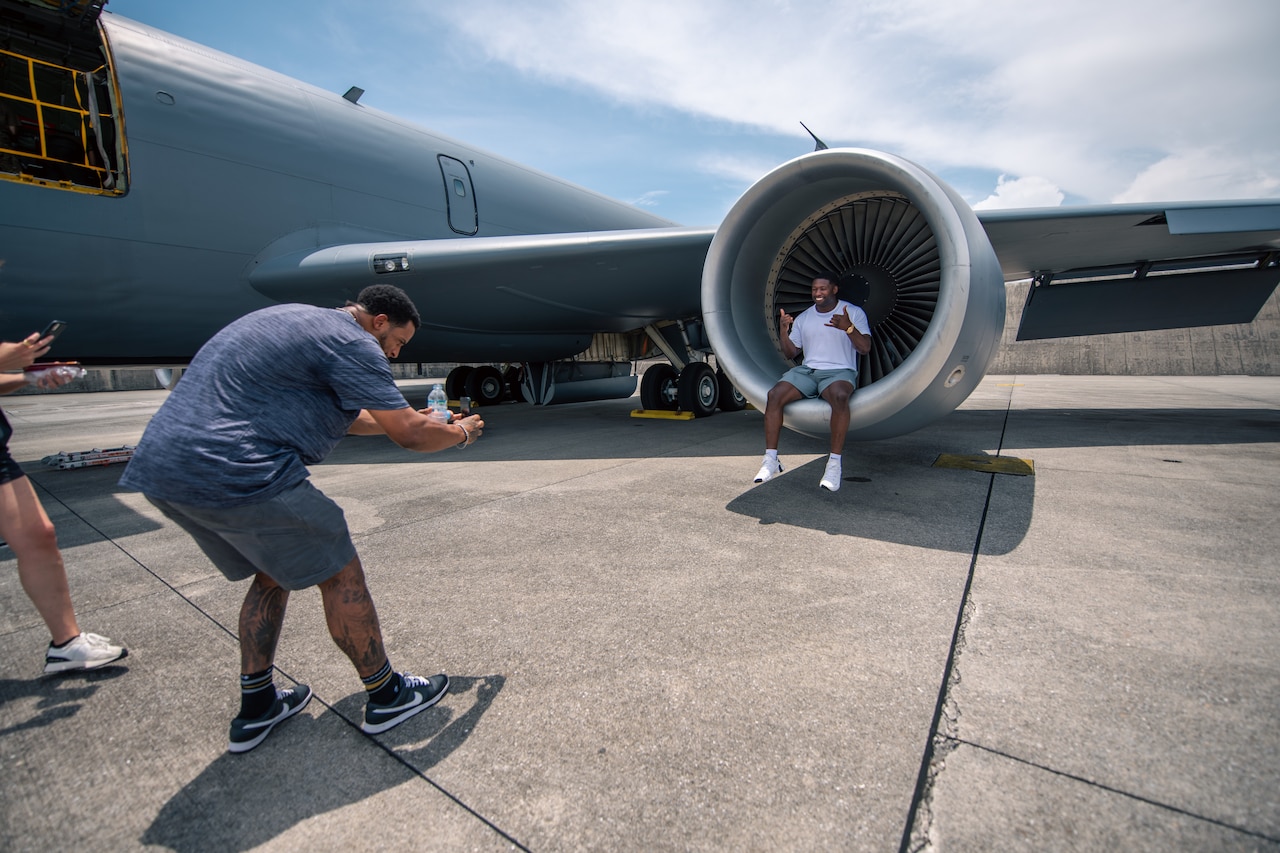 A man takes a photo of another man posing on the ledge of a jet engine.
