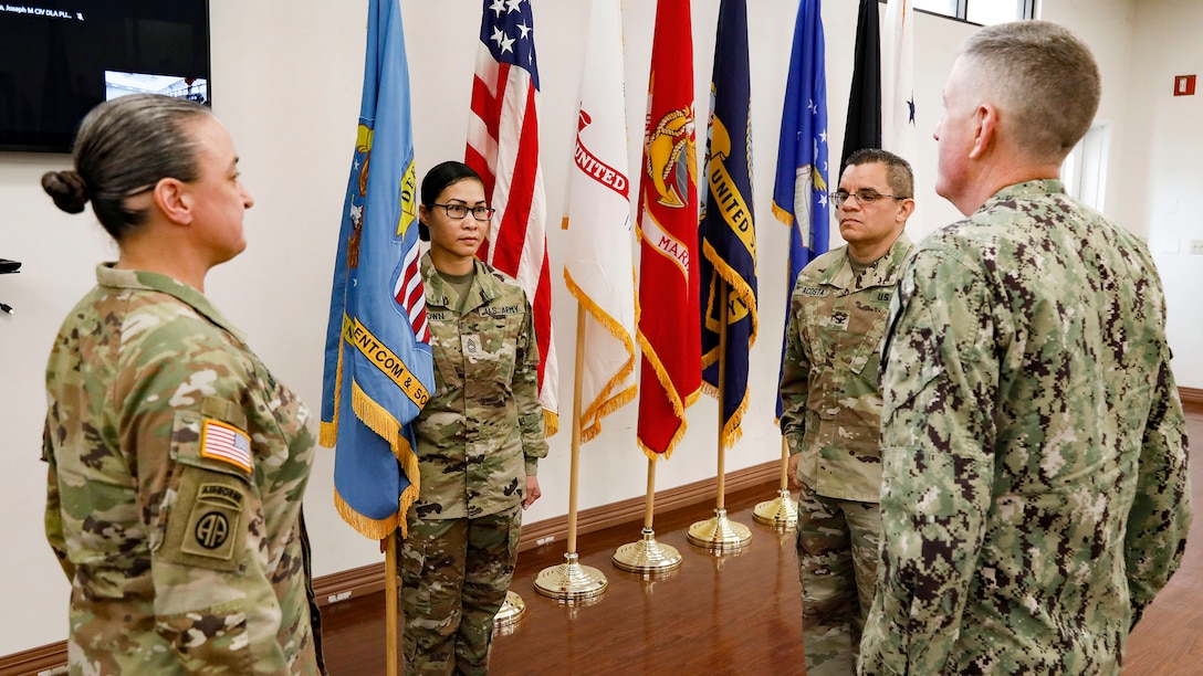 Two women and one man in camo uniforms stand in front of a set of flags facing one another.