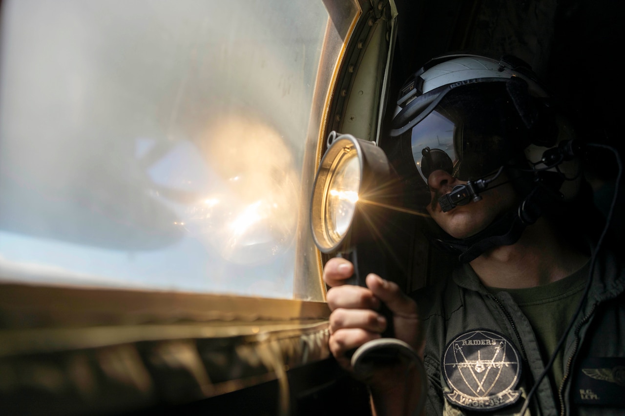 A Marine wearing protective gear shines a light outside of an aircraft’s window.