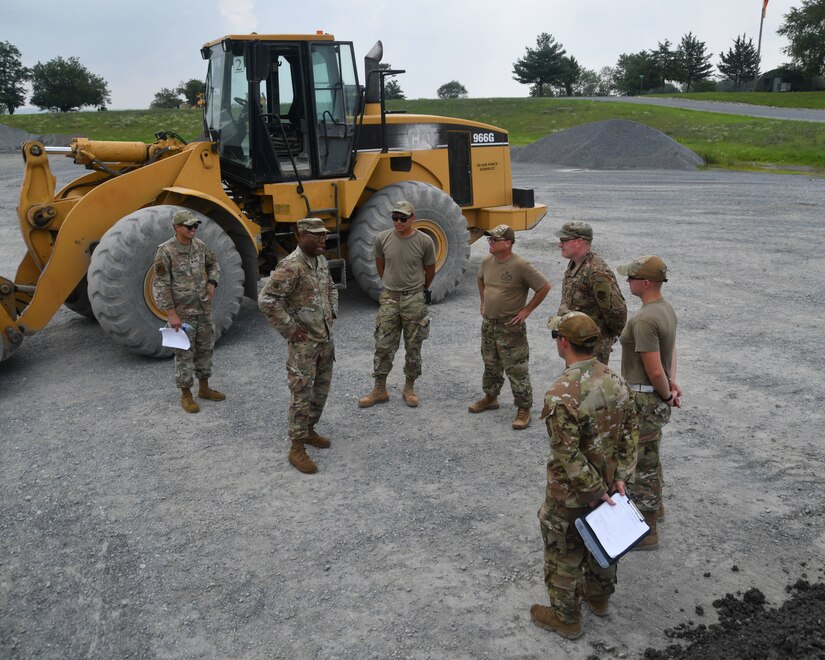 Military members in uniform stand in front of a front end loader.