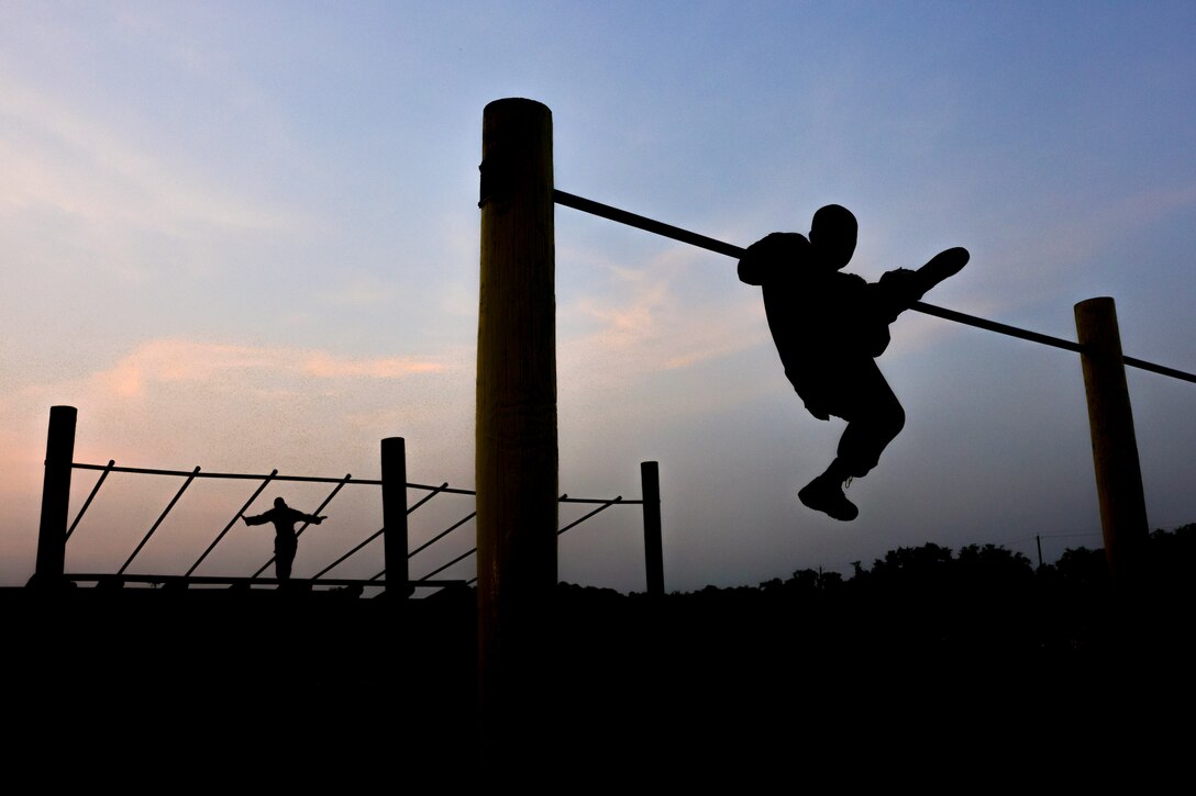 A Marine Corps recruit climbs an elevated bar as another walks across monkey bars in the background as seen in silhouette.