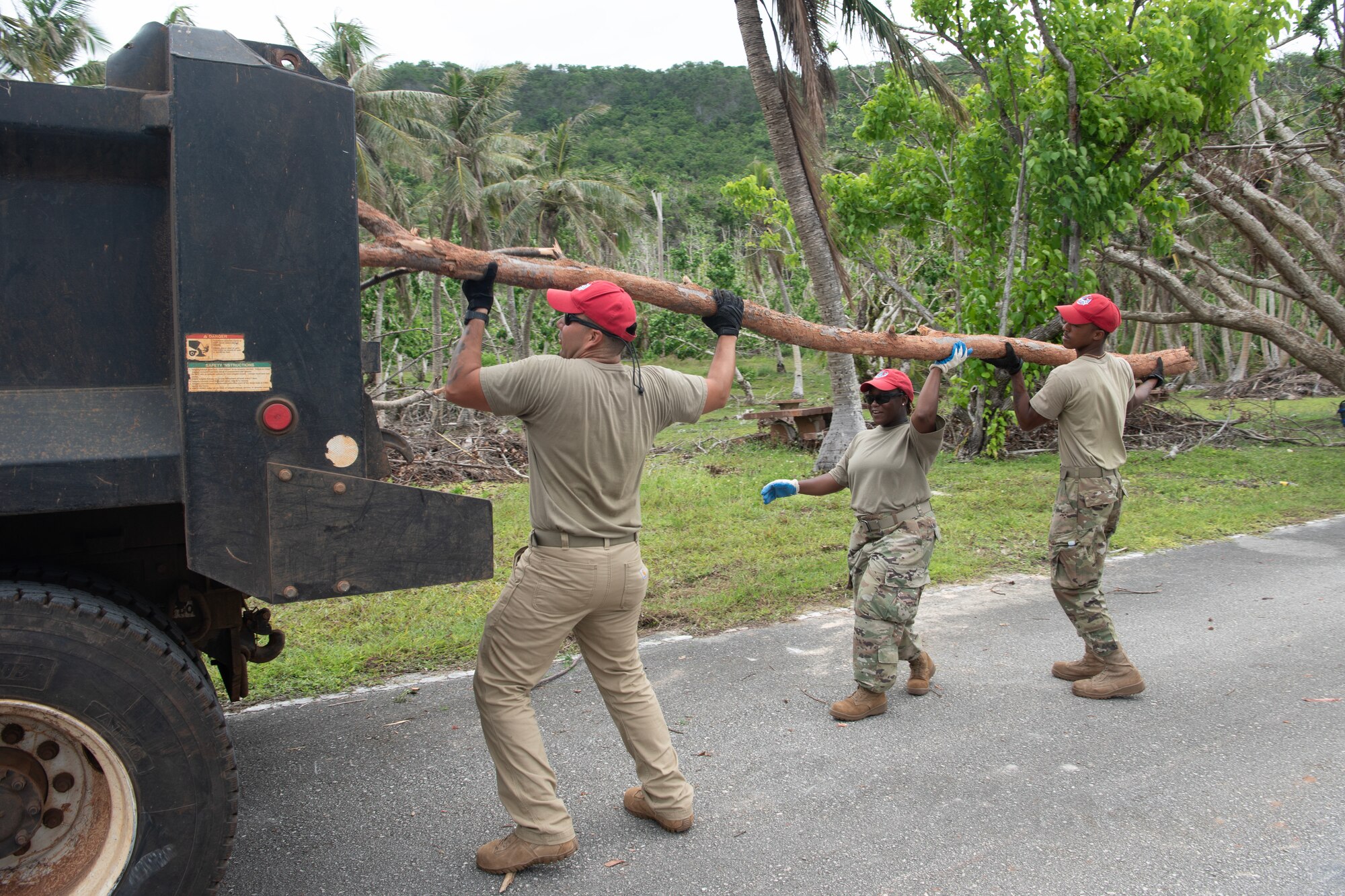 Three Airmen from a visiting squadron lifts a large tree branch.