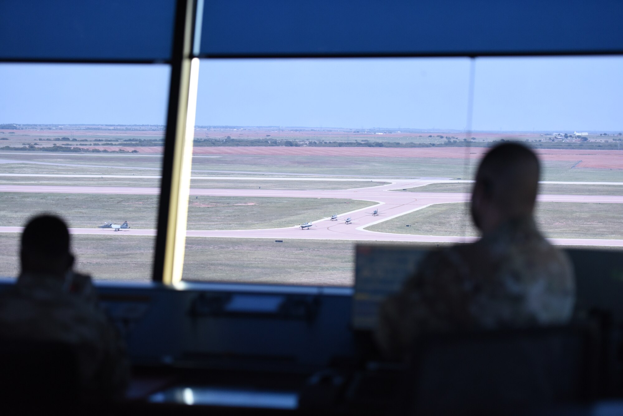 Two blurry figures look out of air traffic control tower window at in-focus jets below