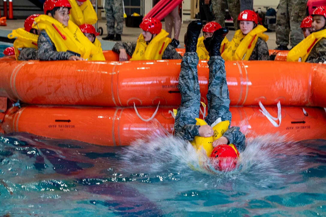 Service members sit in an orange inflatable raft as one plunges into water back first.