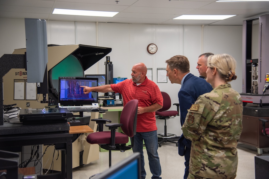 Three men and a woman look at the screen of one of the testing machines in a lab. The woman is wearing a camoflage military uniform.